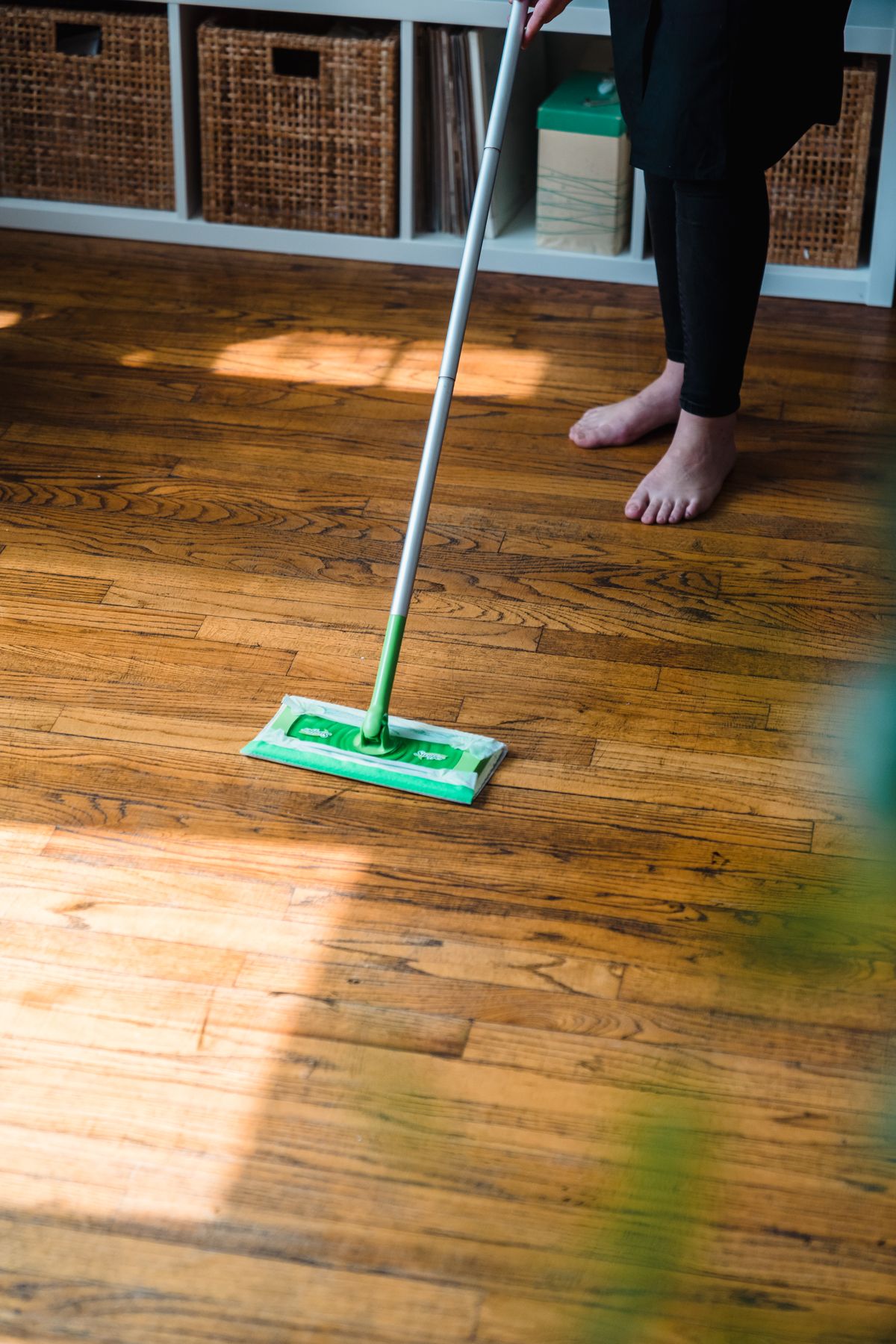 Person using a green flat mop to clean a sunlit wooden floor.