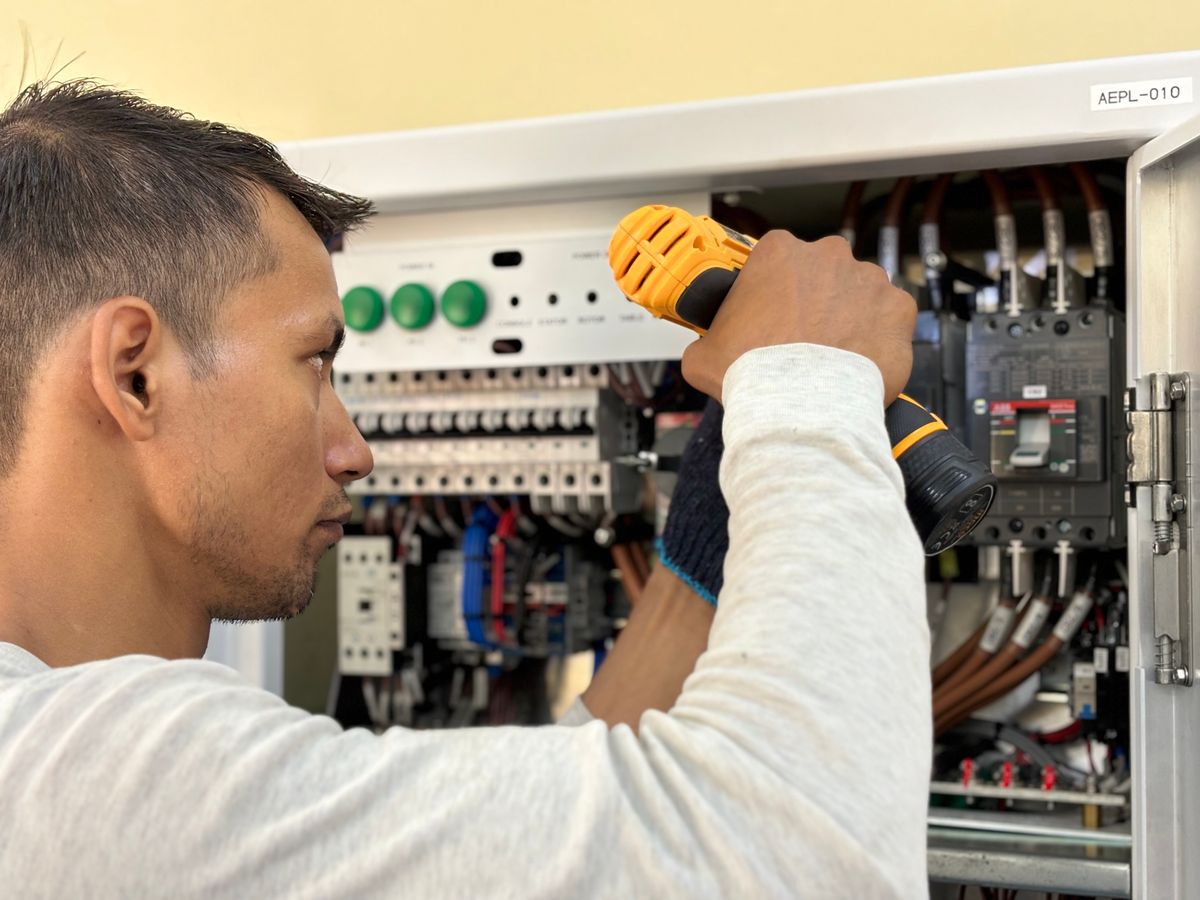 Electrician using a cordless drill at an open electrical control panel.